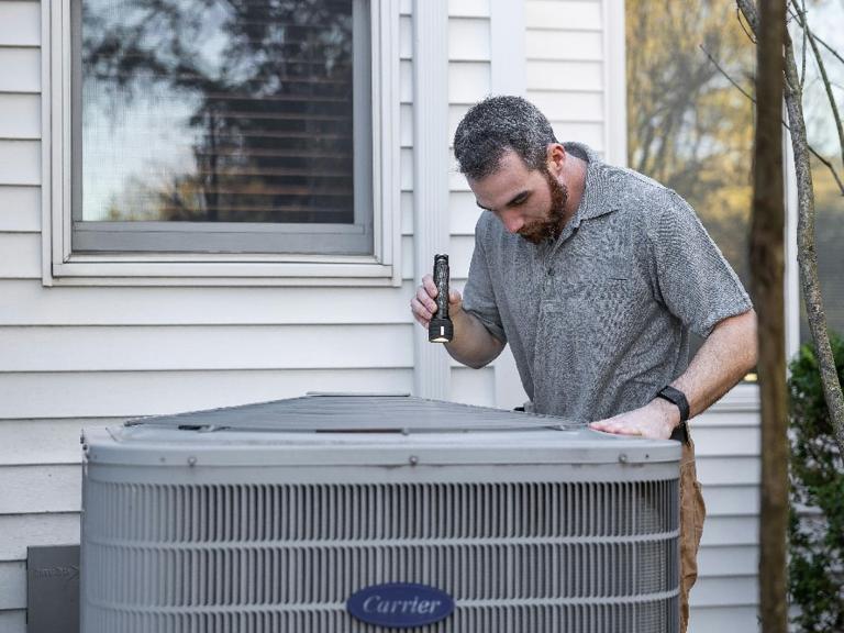 Technician inspecting an outdoor air conditioning unit with a flashlight next to a home window