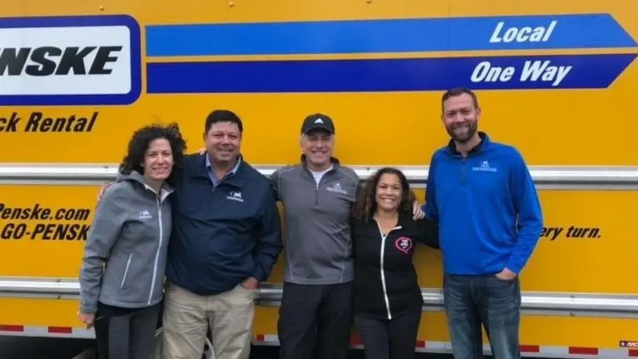 Five people smiling in front of a bright yellow Penske truck with the "Local One Way" logo. They appear friendly and relaxed.